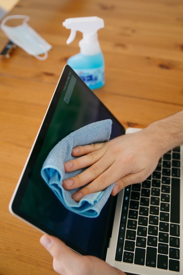 services-02 A person wipes a laptop screen using a blue cloth and spray bottle on a wooden table for hygiene.